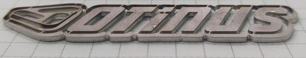Precyzyjnie wycięte laserowo metalowe logo Otinus przedstawiające wysoką jakość i gładkość krawędzi.