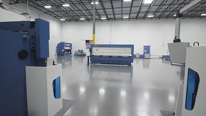 Wnętrze nowoczesnego, przestronnego showroom'u z jasną, wypolerowaną podłogą. Na środku i w tle stoją niebiesko-białe maszyny przemysłowe, rozmieszczone w dużych odstępach. Na ścianie widoczna jest amerykańska flaga, a całość oświetlają rzędy lamp podwieszonych pod wysokim stropem.